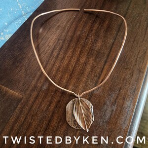 May include: A copper-toned metal collar necklace featuring a circular textured pendant with an attached leaf-shaped charm. The necklace has an open-ended design and the text TWISTEDBYKEN.COM is printed in white at the bottom of the image.