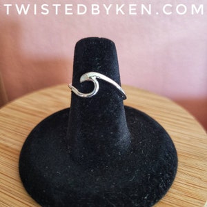 May include: A silver wave ring on a black ring display stand. The ring is a simple design with a wave shape.