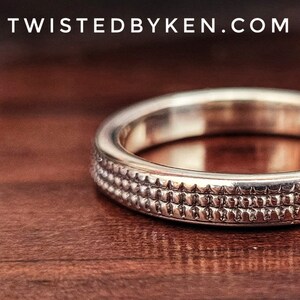 May include: A close-up of a silver wedding band with a textured design. The band is on a wooden surface.