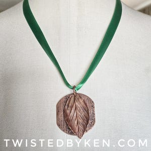 May include: A copper-colored leaf pendant with a detailed leaf design, suspended from a green satin ribbon. The pendant is attached to the ribbon with a small copper ring. The text "TWISTEDBYKEN.COM" is at the bottom of the image.