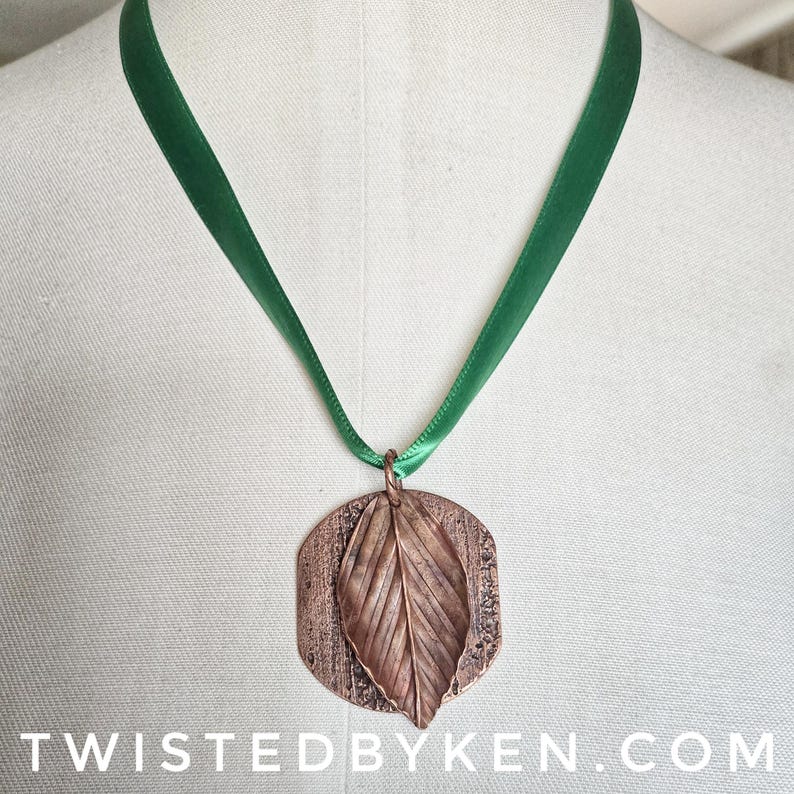 May include: A copper-colored leaf pendant with a detailed leaf design, suspended from a green satin ribbon. The pendant is attached to the ribbon with a small copper ring. The text "TWISTEDBYKEN.COM" is at the bottom of the image.