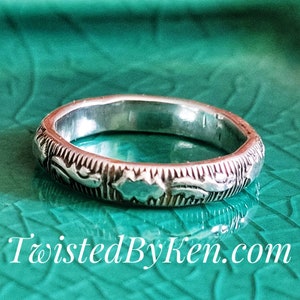 May include: A silver band ring with a delicate floral design. The ring is on a teal green surface.