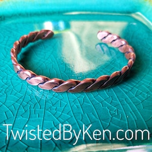 May include: A copper bracelet with a twisted design. The bracelet is on a turquoise background.
