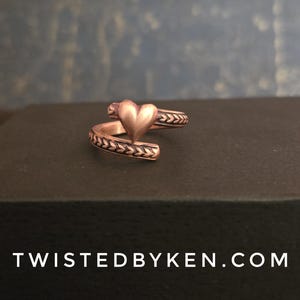 May include: A copper ring with a heart-shaped design and a braided band. The ring is on a brown surface.
