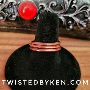 May include: A set of three copper rings stacked on a black display stand. The rings are smooth and have a warm, reddish-brown color.