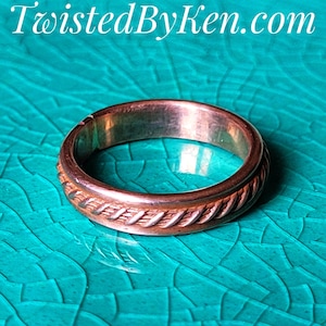 May include: A copper ring with a braided design on the band. The ring is on a turquoise background.