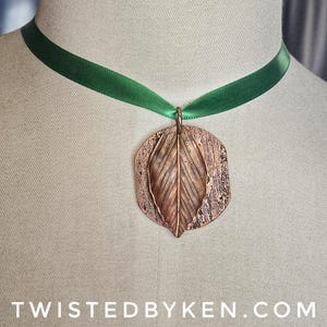 May include: A copper-colored leaf pendant necklace with a textured background, suspended from a green satin ribbon. The pendant features a detailed leaf design. The text "TWISTEDBYKEN.COM" is at the bottom of the image.
