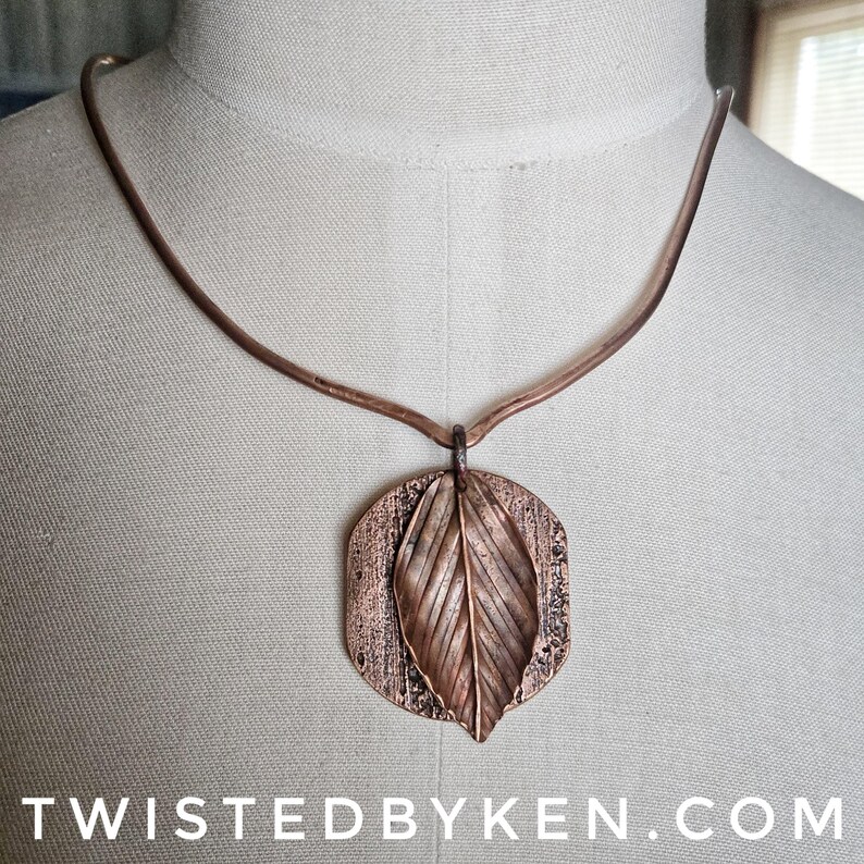May include: A copper-colored necklace featuring a leaf pendant. The pendant has a textured, rectangular base with a detailed leaf overlay. The necklace is displayed on a mannequin. The text "TWISTEDBYKEN.COM" is at the bottom.
