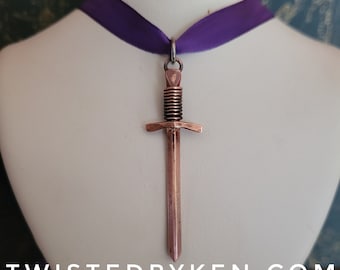May include: A copper sword pendant necklace with a purple ribbon. The sword has a detailed hilt and a pointed tip. The pendant is hanging from a silver chain.