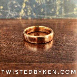 May include: A simple gold band ring, with a smooth, polished finish. The ring is sitting on a wooden surface.