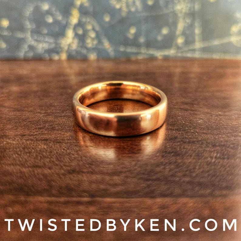 May include: A simple gold band ring, with a smooth, polished finish. The ring is sitting on a wooden surface.