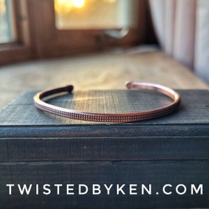 May include: A copper cuff bracelet with a textured design. The bracelet is on a dark wooden surface.