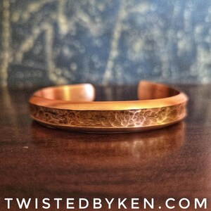 Puede incluir: Un brazalete de cobre martillado con un acabado liso y pulido. El brazalete est&aacute; abierto y tiene una forma ligeramente curvada.