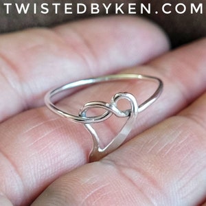May include: A silver ring with a heart-shaped design. The ring is made of a thin band of metal and the heart is formed by a knot in the band.