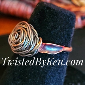 Puede incluir: Anillo artesanal con un diseño de rosa de alambre. La rosa está hecha de alambre plateado y la banda es de color cobre. El anillo tiene una apariencia artística única. El texto "TwistedByKen.com" es visible.