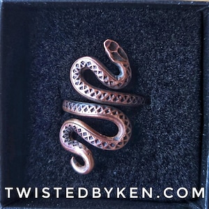 May include: A copper-toned metal ring shaped like a coiled snake with a textured, cross-hatched pattern along its body. The ring is displayed inside a black box with the text TWISTEDBYKEN.COM printed in white at the bottom of the image.