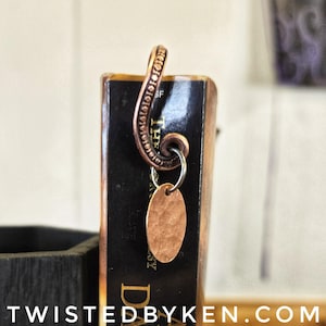May include: A copper bookmark with a decorative swirl design and a hammered copper oval charm. The bookmark is attached to a book with the text "The Da" visible.