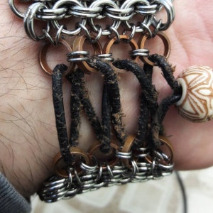 May include: A silver chainmail bracelet with copper rings and black leather straps. The bracelet has a white ceramic bead with a floral design.