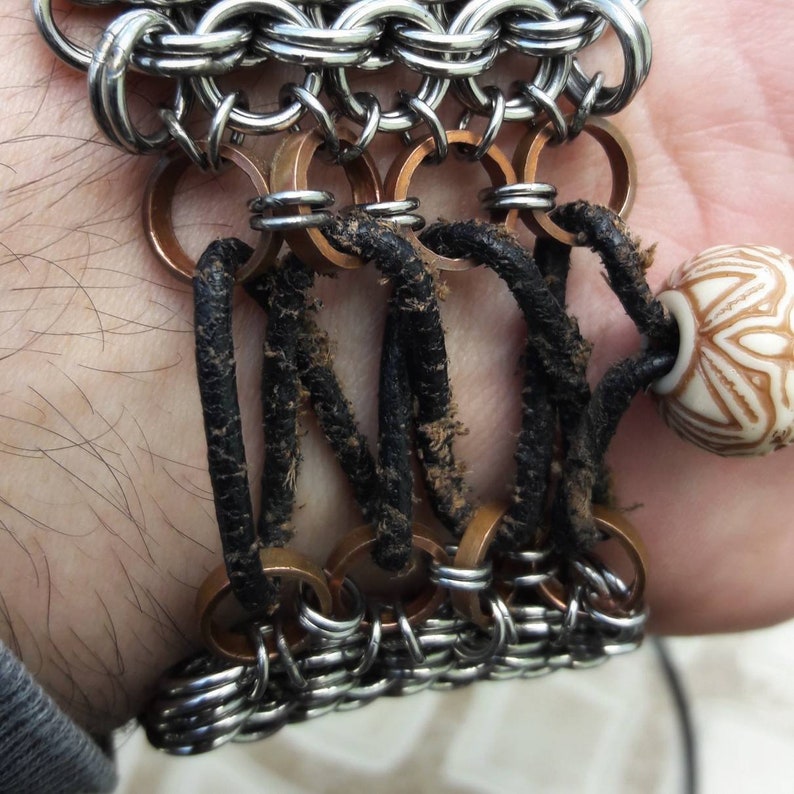 May include: A silver chainmail bracelet with copper rings and black leather straps. The bracelet has a white ceramic bead with a floral design.