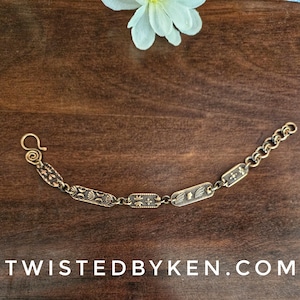 May include: A gold-toned bracelet with a chain link design and rectangular charms featuring floral patterns. The bracelet has a hook clasp.