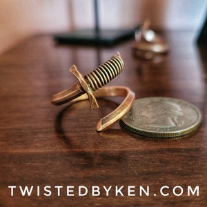 May include: A gold-toned metal ring with a coiled sword design. The ring is lying on a wooden surface next to a US quarter.