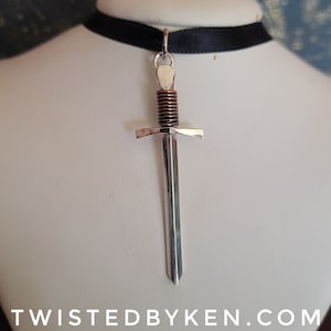 May include: A silver sword pendant with a copper-colored hilt, hanging from a black ribbon choker.