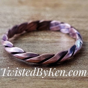 May include: A close-up of a copper ring with a twisted design. The ring is on a wooden surface.