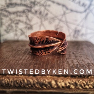 May include: A copper ring with a textured leaf design. The ring is open and has a twisted look. The ring is on a wooden surface with the text "TWISTEDBYKEN.COM" visible.