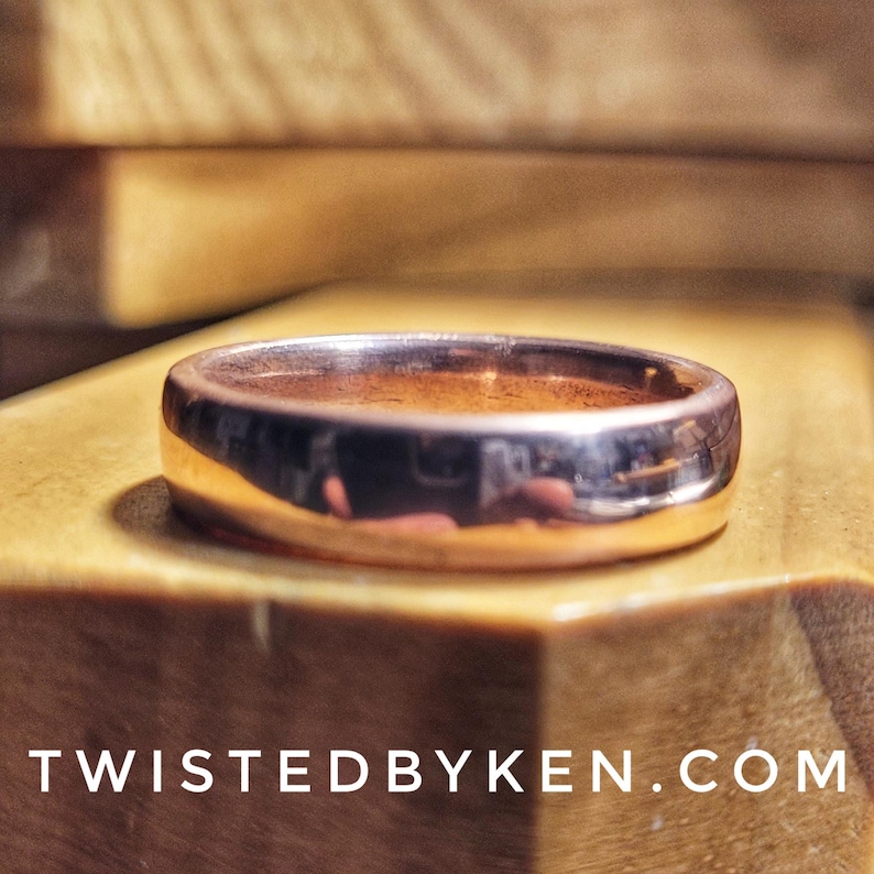 May include: A polished, copper-colored ring with a rounded, smooth surface. The ring is sitting on a wooden surface.