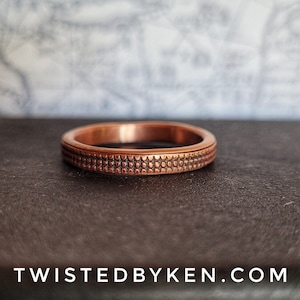 May include: A copper ring with a textured pattern. The ring is on a dark brown surface.