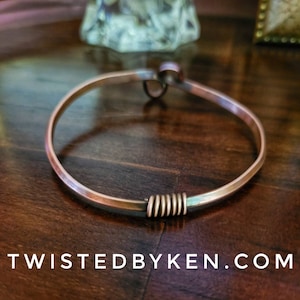 May include: A copper bangle bracelet with a twisted wire detail. The bracelet is on a wooden surface.