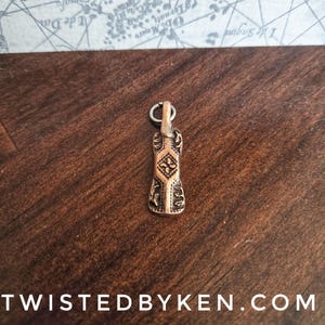 May include: A copper-toned metal pendant featuring an elongated rectangular shape with rounded edges. The surface displays an embossed geometric diamond pattern in the center, flanked by intricate leaf-like motifs on both sides, and includes a small silver-toned jump ring at the top for attachment.
