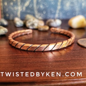 May include: A copper bracelet with a twisted design. The bracelet is made of a single piece of copper that has been hammered and twisted to create a unique and textured look.