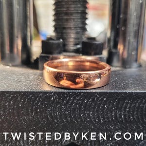 May include: A close-up of a copper ring, polished to a high shine, resting on a black surface. The ring is thick and has a rounded profile. The reflection of the surrounding environment is visible in the ring's surface.