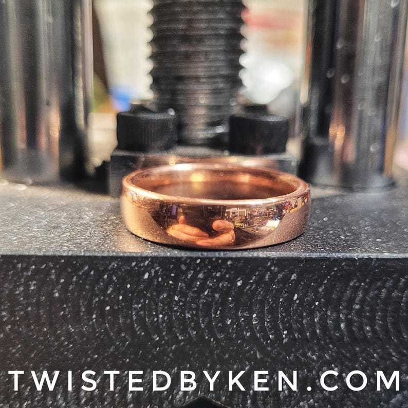 May include: A close-up of a copper ring, polished to a high shine, resting on a black surface. The ring is thick and has a rounded profile. The reflection of the surrounding environment is visible in the ring's surface.