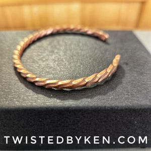 May include: A copper bracelet with a twisted, textured design. The bracelet is made of a single piece of copper that has been hammered and twisted to create a unique and eye-catching look.