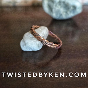 May include: A copper-colored ring with a twisted rope design rests on a small, light-colored stone. The ring is the focal point, showcasing its intricate texture and metallic sheen. The website address "TWISTEDBYKEN.COM" is displayed at the bottom of the image.
