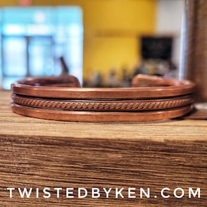 May include: A copper bracelet with a braided design. The bracelet is on a wooden surface.