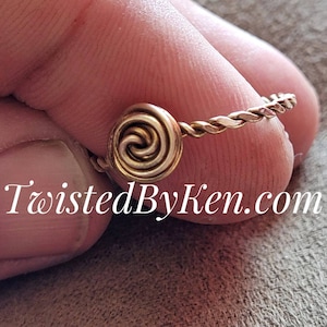 May include: A gold-toned ring with a twisted wire band and a spiral design in the center. The ring is on a finger.