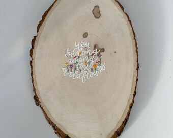 May include: An oval-shaped natural wood slice with bark edges featuring the hand-painted white cursive text "you belong among the wildflowers" surrounded by small, colorful painted flowers in shades of yellow, orange, purple, and green.