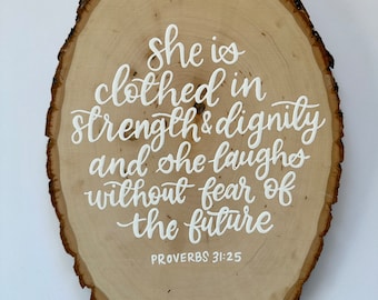 May include: A wooden plaque with a natural bark edge. The plaque features white cursive text that reads "she is clothed in strength & dignity and she laughs without fear of the future" with "PROVERBS 31:25" below.