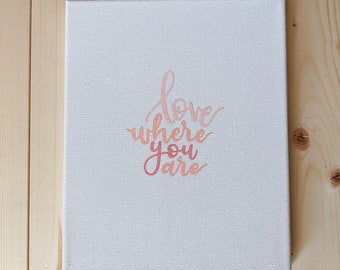 May include: A white canvas with the phrase "love where you are" written in pink and orange lettering.