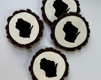 May include: Four circular wood slice coasters with natural dark brown bark edges. Each coaster features a smooth, cream-colored painted center with a solid black silhouette of the state of Wisconsin centered on the surface of the wood.