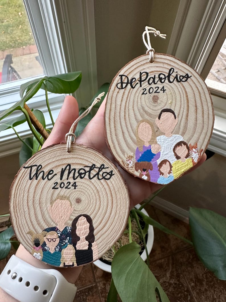May include: Two round wooden ornaments with painted family portraits and the family names "The Motts" and "DePaolis" with the year 2024.