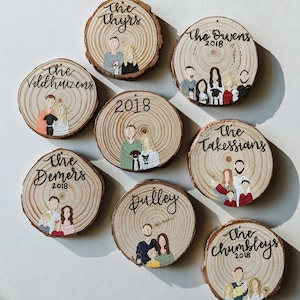 May include: Eight round wood slices with painted family portraits and names. Each slice has a white background with brown wood grain showing through. The names are written in black cursive script and the year 2018 is included on each slice. The portraits are painted in a cartoon style with simple features and bright colors.