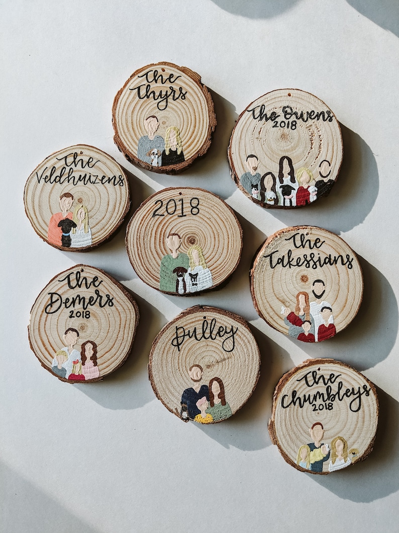 May include: Eight round wood slices with painted family portraits and names. Each slice has a white background with brown wood grain showing through. The names are written in black cursive script and the year 2018 is included on each slice. The portraits are painted in a cartoon style with simple features and bright colors.