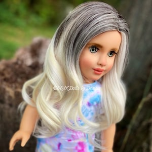May include: A doll with long, wavy, blonde hair with grey streaks. The doll is wearing a blue and white tie-dye shirt. The doll has blue eyes and is looking to the right.