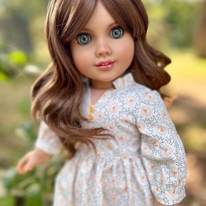 May include: A doll with long brown wavy hair, wearing a white dress with a floral pattern and green eyes.
