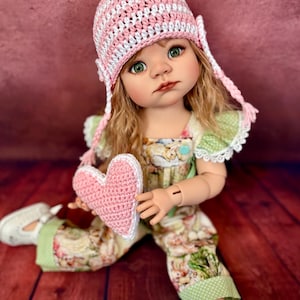 May include: A doll wearing a pink and white striped crochet hat with matching braids and a pink heart. The doll is dressed in a floral print jumpsuit with green accents and white shoes. The doll has green eyes and blonde hair.
