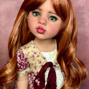 May include: A doll with auburn hair and green eyes. The doll has freckles and is wearing a floral dress with a burgundy ribbon. The doll's hair is styled in loose waves.